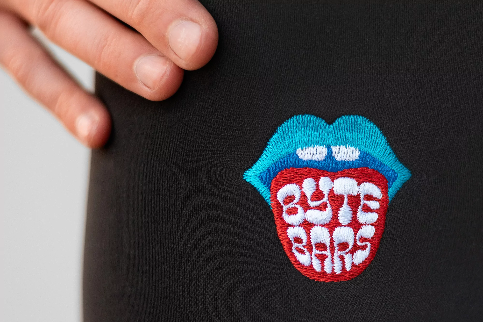 Close-up of a black garment featuring an embroidered patch of a mouth with text 'BYTE BARS', accented by a person's fingers.