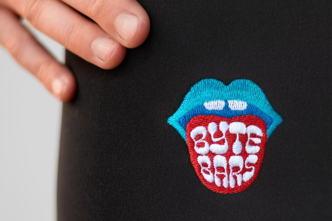 Close-up of a black garment featuring an embroidered patch of a mouth with text 'BYTE BARS', accented by a person's fingers.