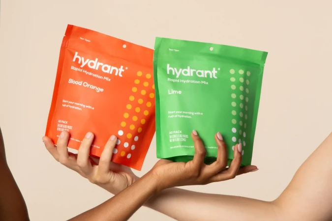 Two hands holding bright orange and green Hydrant rapid hydration mix pouches against a plain light background