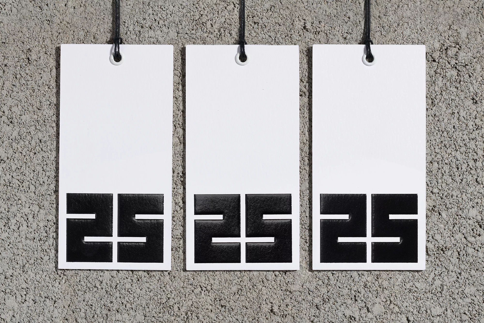 Three white tags with black '25' graphic hang against a textured grey wall, displayed in a simple, eye-level, studio composition.