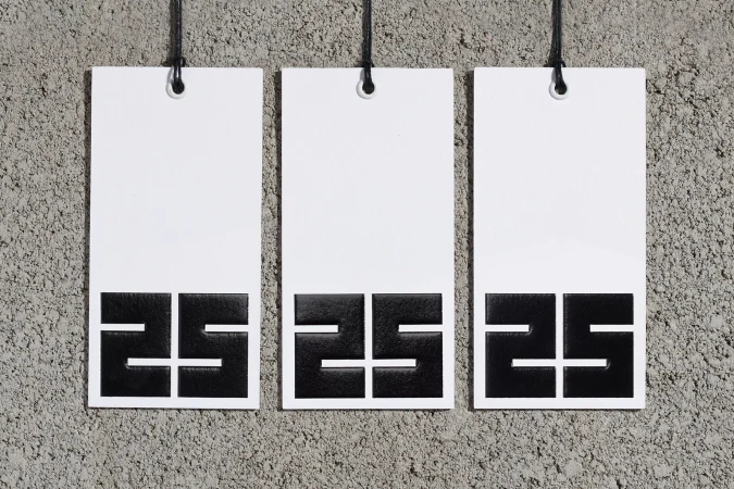 Three white tags with black '25' graphic hang against a textured grey wall, displayed in a simple, eye-level, studio composition.