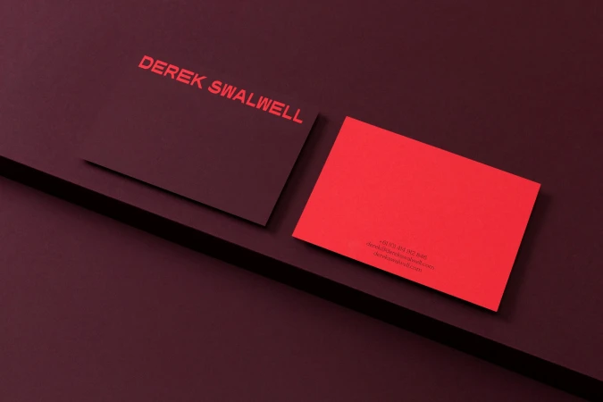 Two business cards, one dark red with red text and the other bright red, rest on a dark red surface with sharp angles.