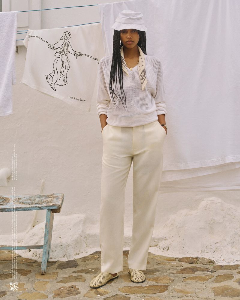 A woman in a white outfit stands on a stone surface with a stool and a drying rack in the background, posing for a fashion shot.
