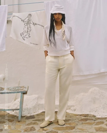 A woman in a white outfit stands on a stone surface with a stool and a drying rack in the background, posing for a fashion shot.