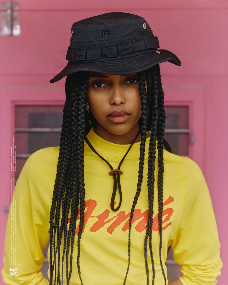 A young woman with long braids poses against a pink wall, wearing a yellow shirt and black bucket hat, in a medium portrait shot.