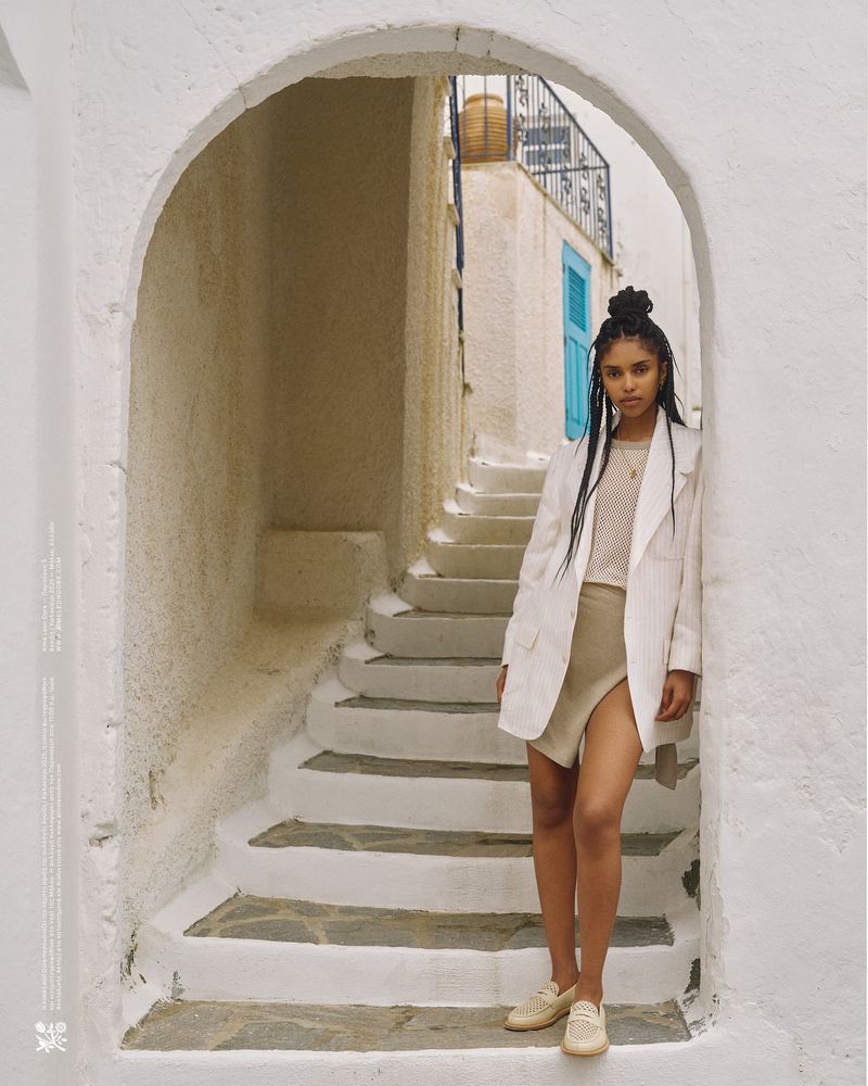 A woman in a blazer and shorts poses within an arched whitewashed passage, bathed in natural light on a sun-drenched day.