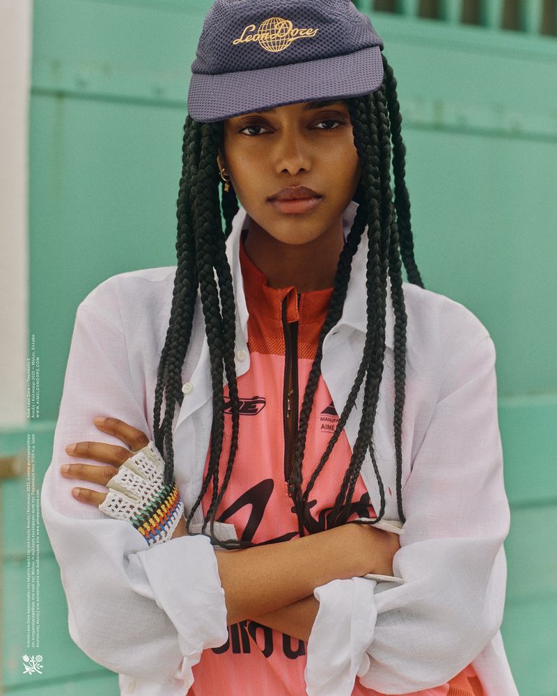 Stylish portrait of a young woman with braids wearing a cap and white shirt, arms crossed, against a teal wall in natural light.