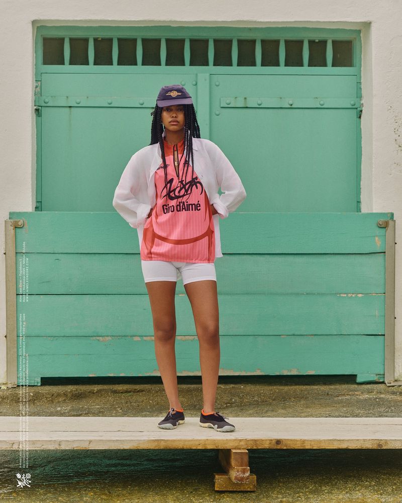 Young adult woman in streetwear stands in front of a teal building in an outdoor setting. She wears a baseball cap, shorts, and sneakers.