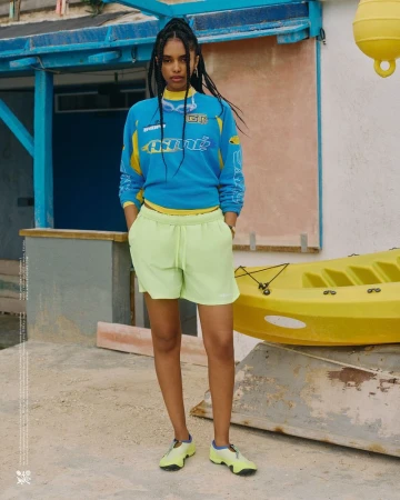 An adult woman poses outdoors wearing a blue and yellow sweatshirt and lime green shorts and athletic shoes. A yellow kayak is visible.