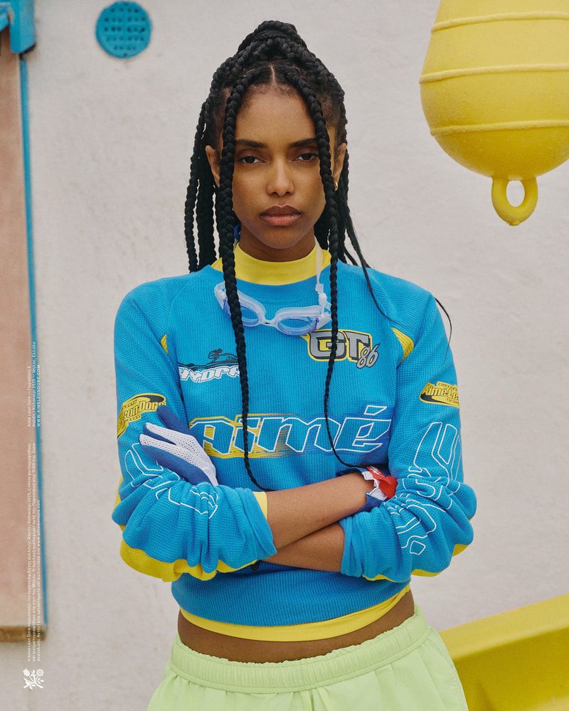 A young Black woman with braided hair poses indoors wearing a turquoise sweater, lime-green shorts, and goggles around her neck.