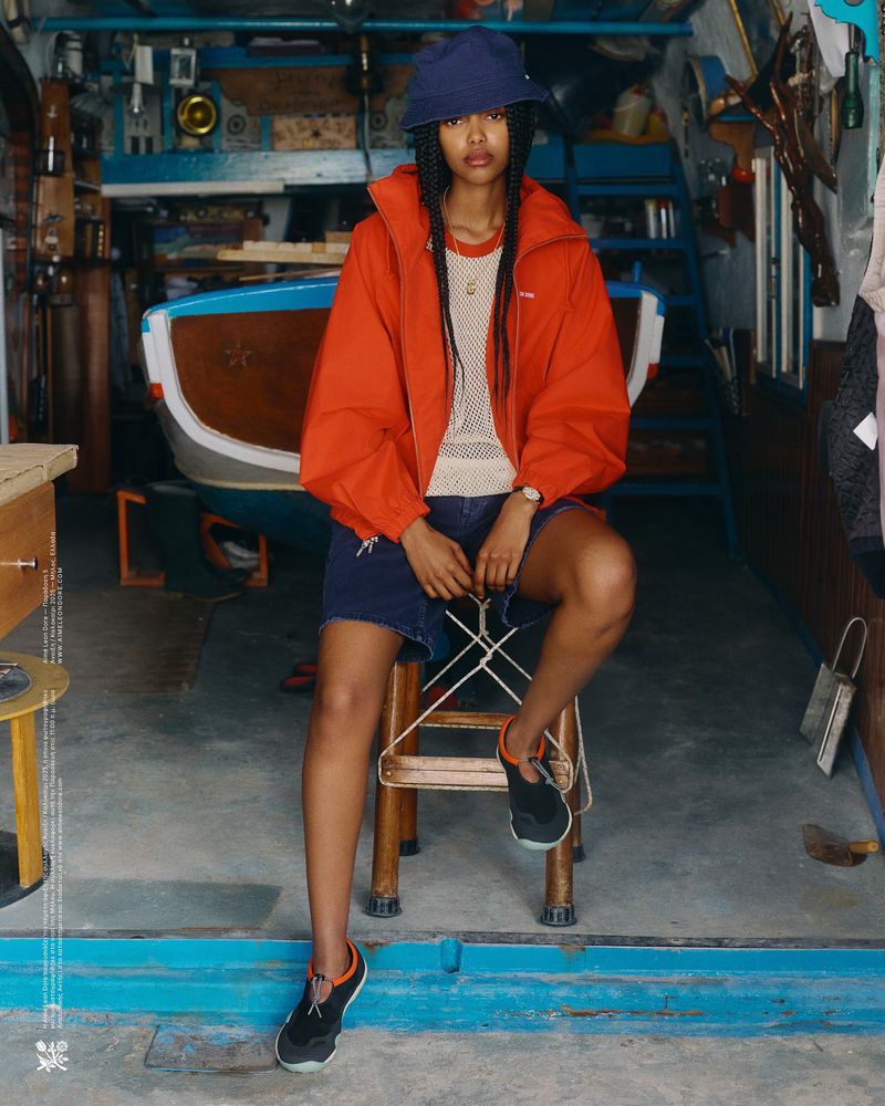 Adult woman in orange jacket and bucket hat seated indoors in front of small boat with blue accents and neutral interior details.