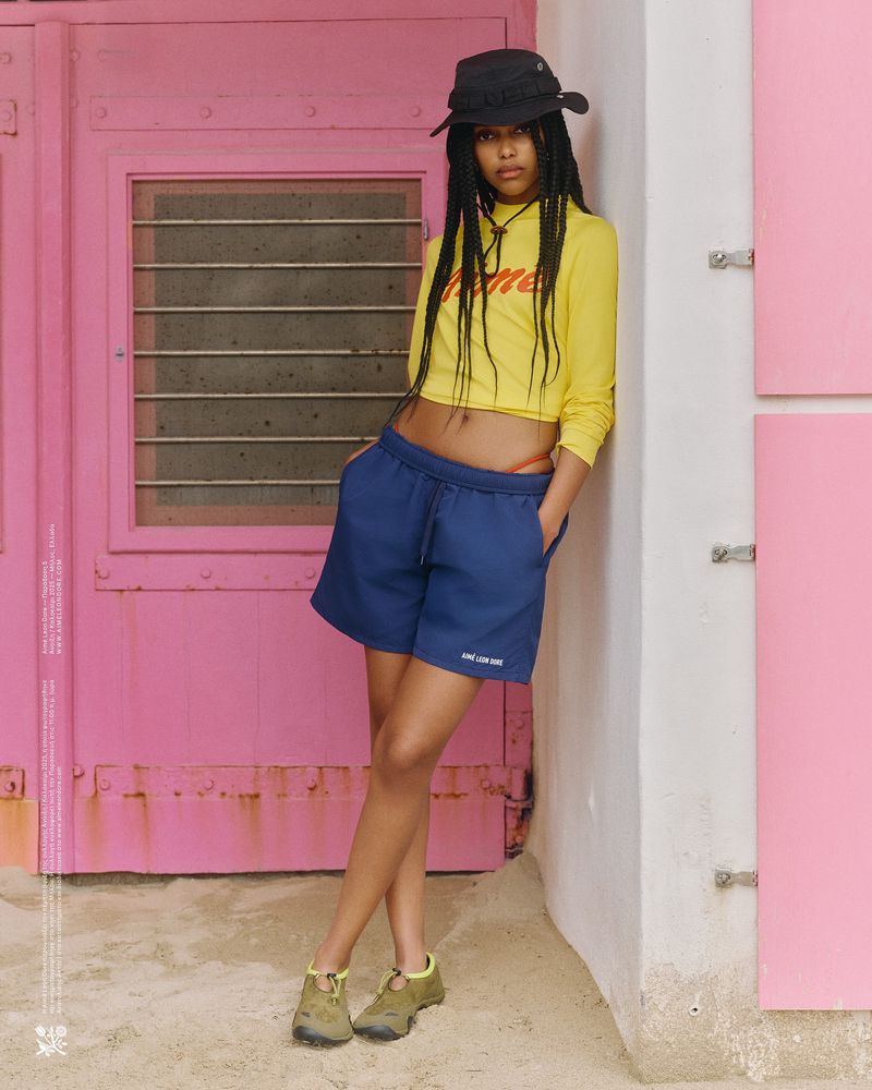 A young adult female poses outdoors in a yellow crop top, blue shorts, and a hat with braided hair, leaning against a pink wall.