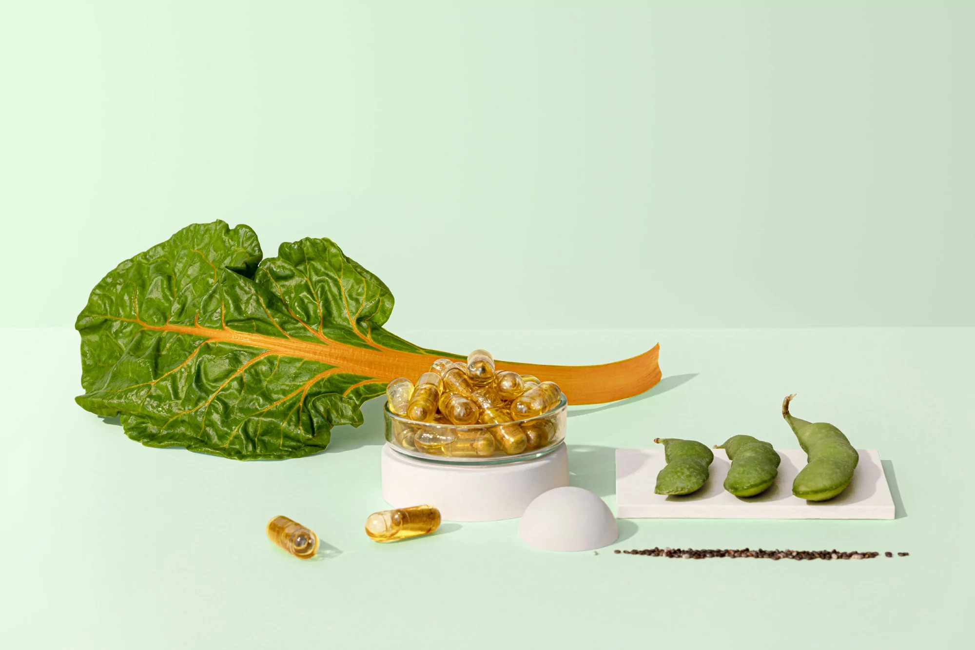 A vibrant still life featuring a chard leaf, capsules in a glass dish, and three edamame pods on a light green background.
