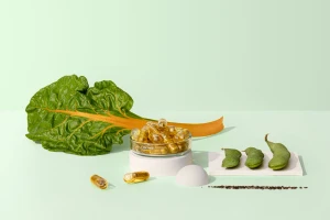 Clear glass petri dish filled with golden gel capsules on a white pedestal alongside green chard and pea pods.