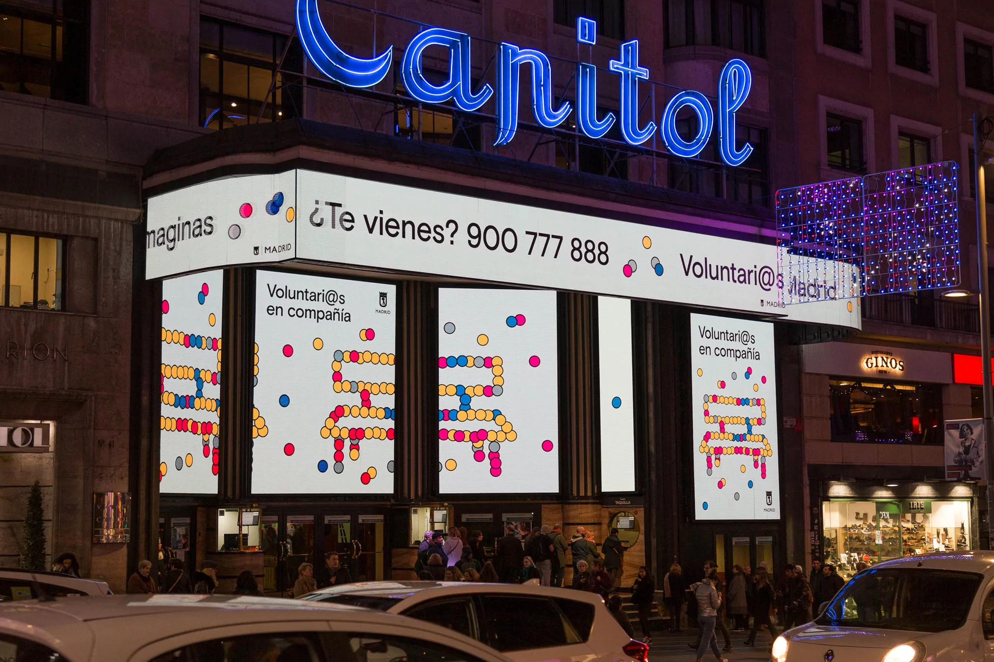 Capitol building in Madrid featuring a volunteer recruitment advertisement on a digital display, illuminated by street and neon lighting.