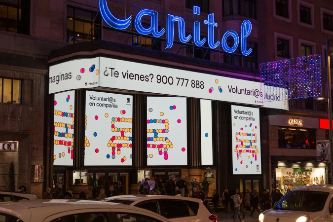 Capitol building in Madrid featuring a volunteer recruitment advertisement on a digital display, illuminated by street and neon lighting.