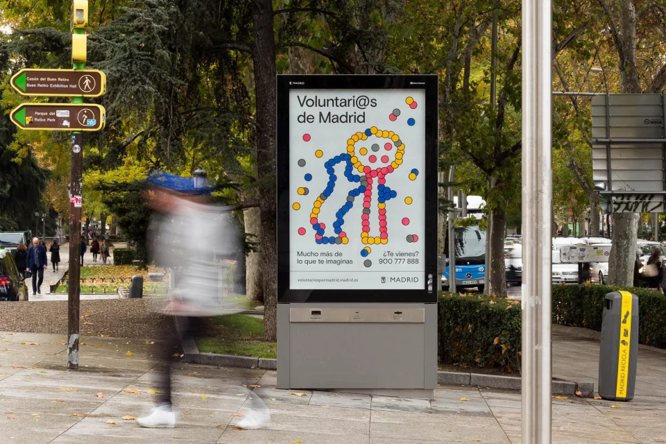 Outdoor digital billboard featuring colorful illustration for Madrid volunteers, set on a city street with blurred pedestrian and trees.