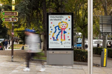 Digital billboard displaying Voluntarios de Madrid graphic identity on a city street with blurred pedestrian passing