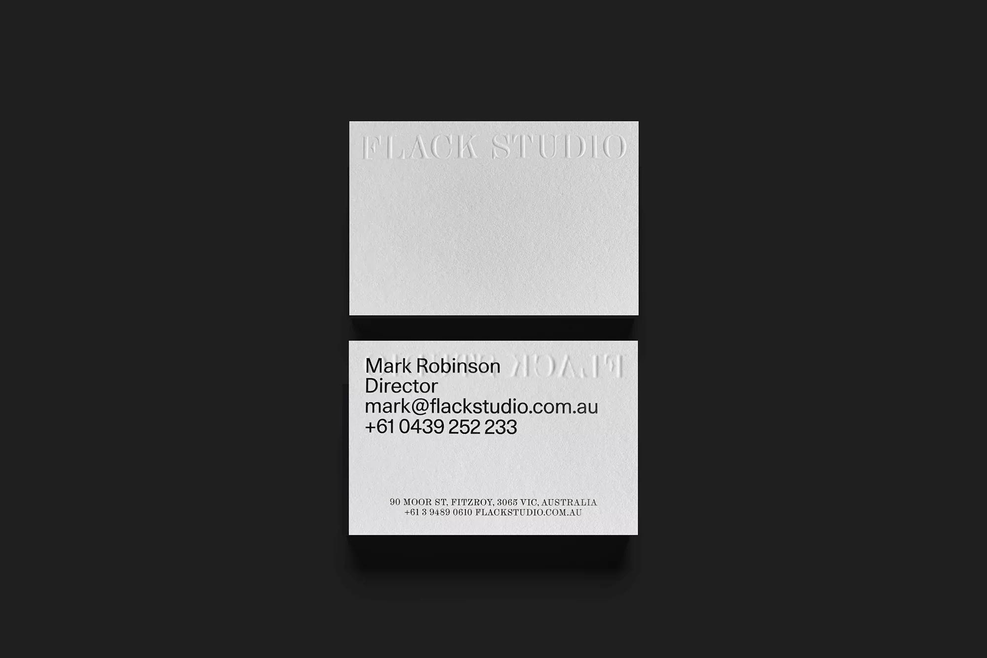 Two white business cards on a dark gray surface. The cards feature the embossed text 'FLACK STUDIO' and contact information.