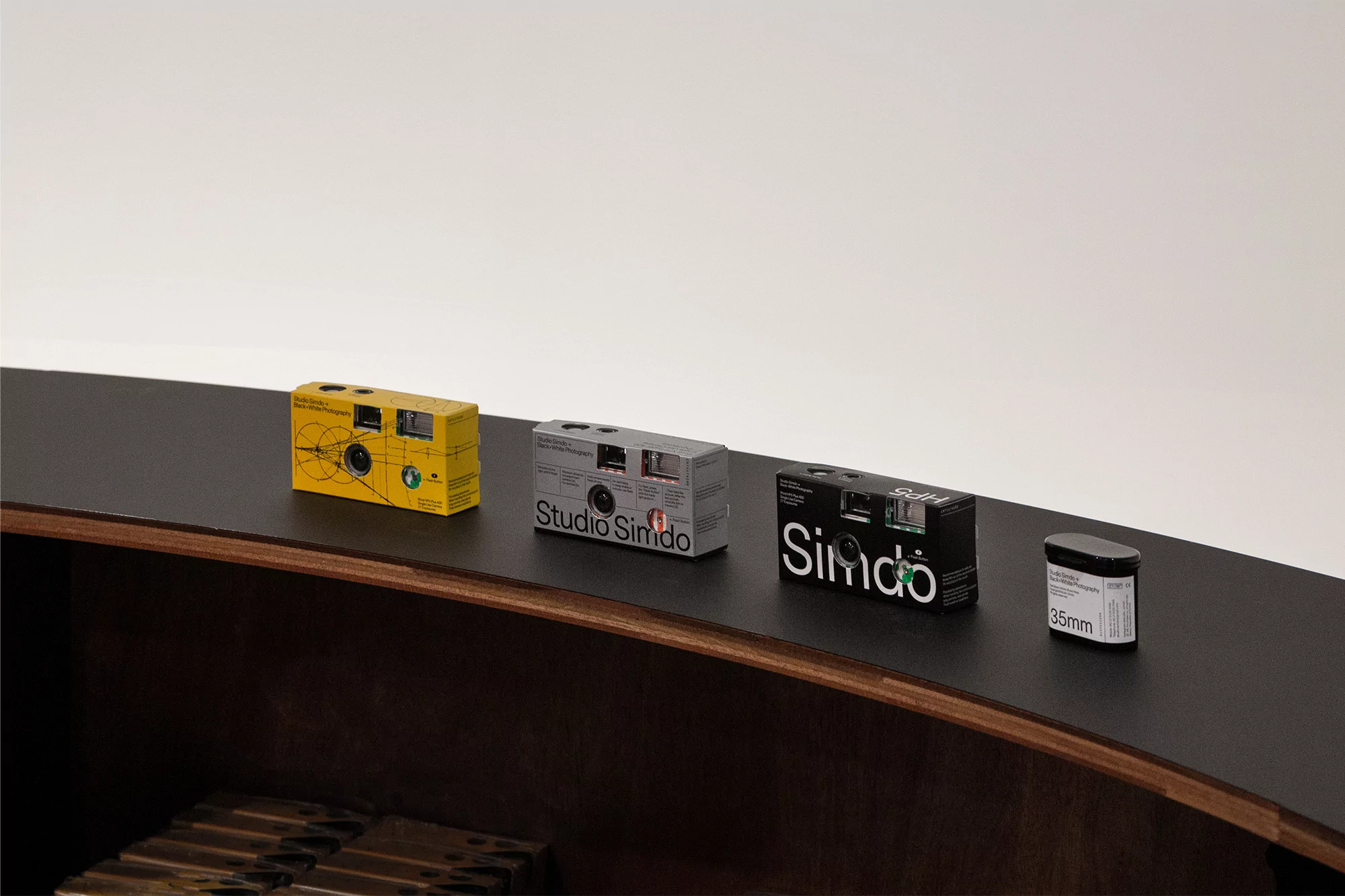 Disposable cameras and a roll of film are arranged on a sleek, curved dark wooden surface against a minimalist white backdrop in a studio setting.