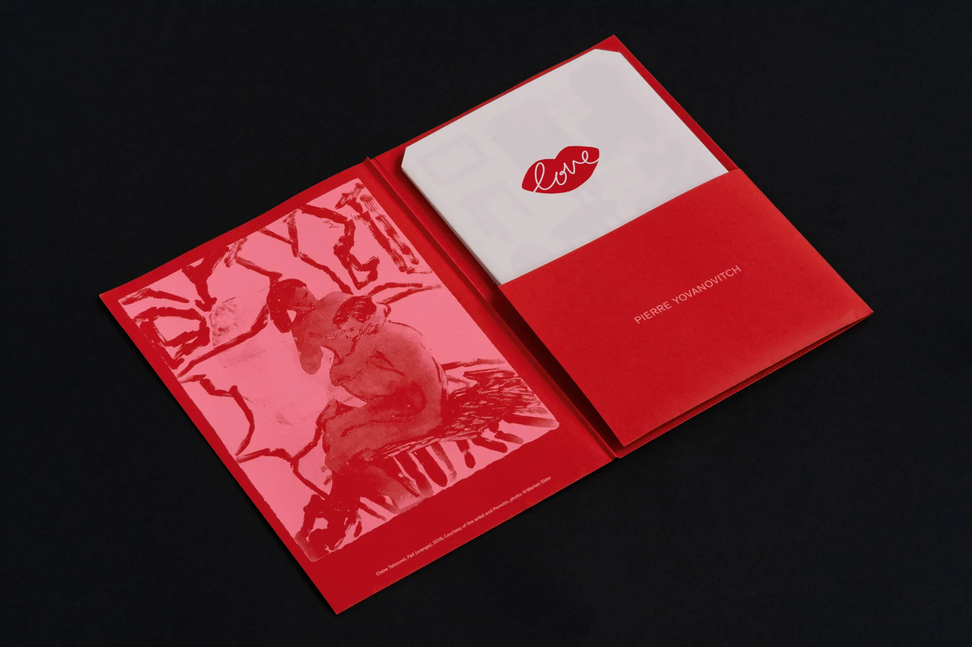 Red folder with art print featuring embracing figures and a card with the word 'love' and lip icon on a stark black surface.