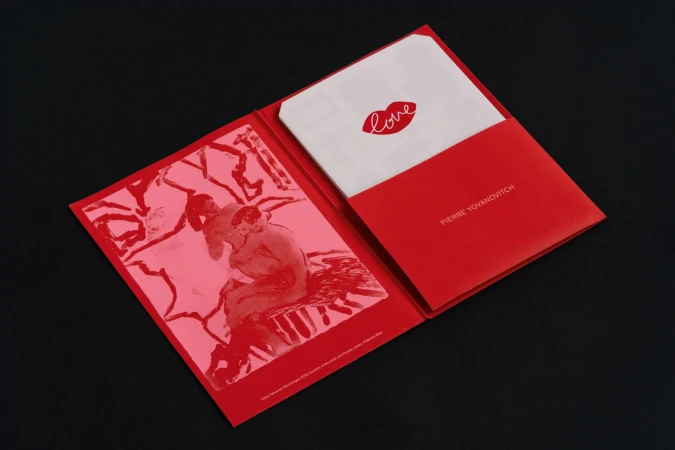Red folder with art print featuring embracing figures and a card with the word 'love' and lip icon on a stark black surface.