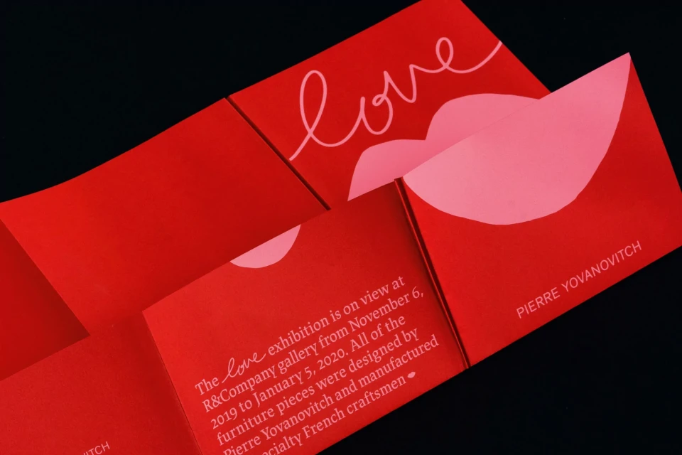 Close-up of a vibrant red exhibition booklet featuring a stylized graphic of the word “love” with a lip shape, set against a black backdrop.