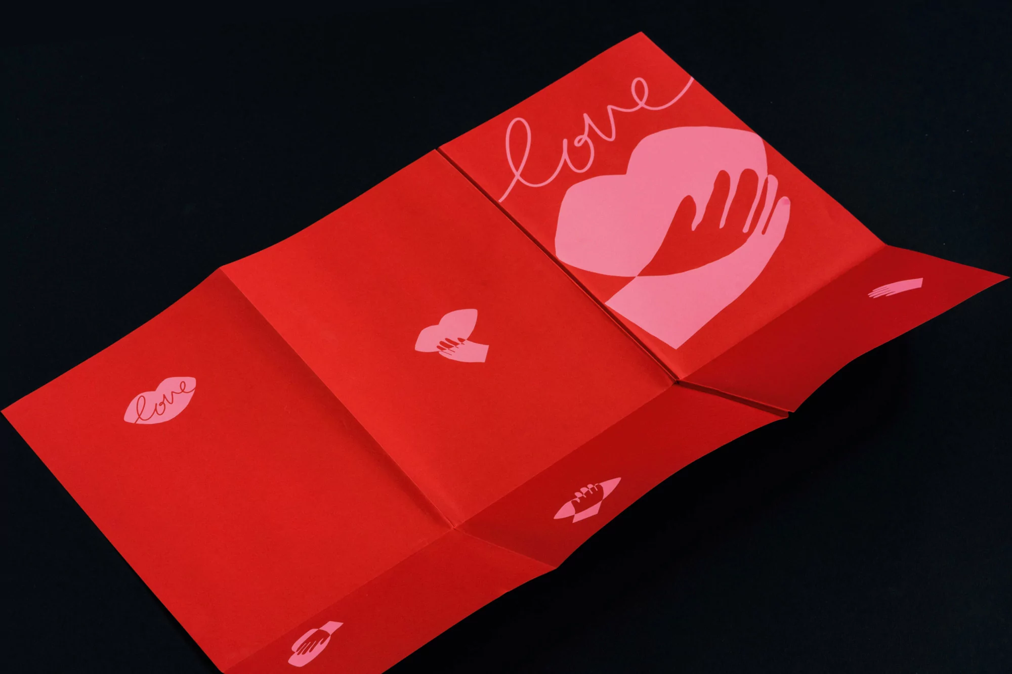 Red folded paper with pink graphics, including a heart held by hands and the word 'love,' on a black background. Valentine's Day design.