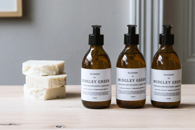 Three amber glass pump bottles of lavender and patchouli liquid soap on a wooden table, next to a stack of three bars of soap.