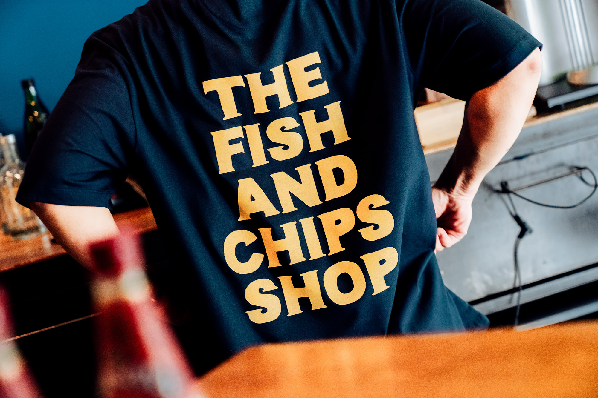 Person wearing a black t-shirt with a yellow 'The Fish and Chips Shop' print, standing indoors with bottles in the background.