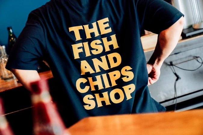 Person wearing a black t-shirt with a yellow 'The Fish and Chips Shop' print, standing indoors with bottles in the background.
