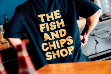 Person wearing a dark blue t-shirt with large yellow text reading THE FISH AND CHIPS SHOP on the back