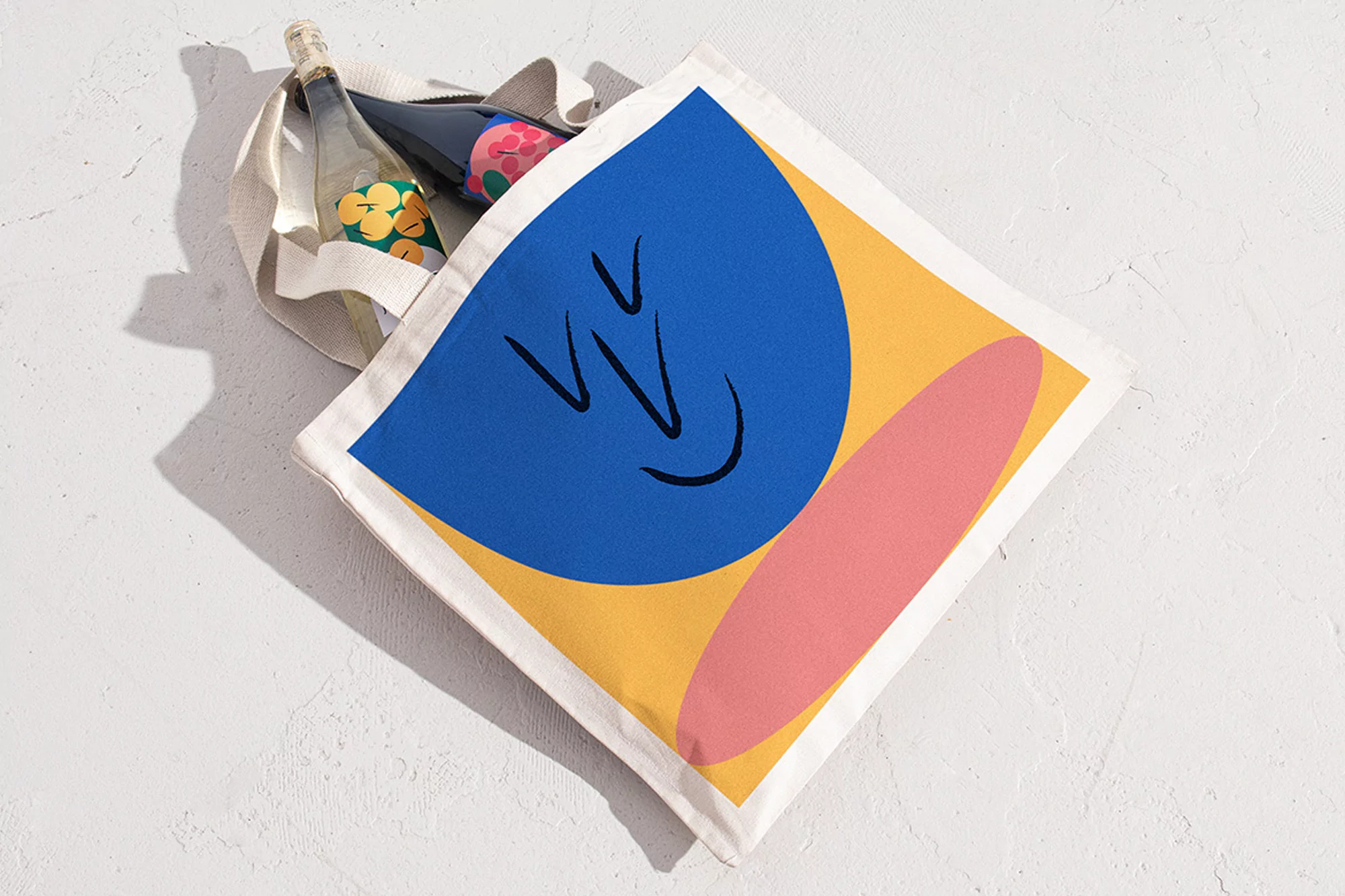 A canvas tote bag with a stylized face design, two glass bottles inside, and white textured background. Neutral and bold color palette.