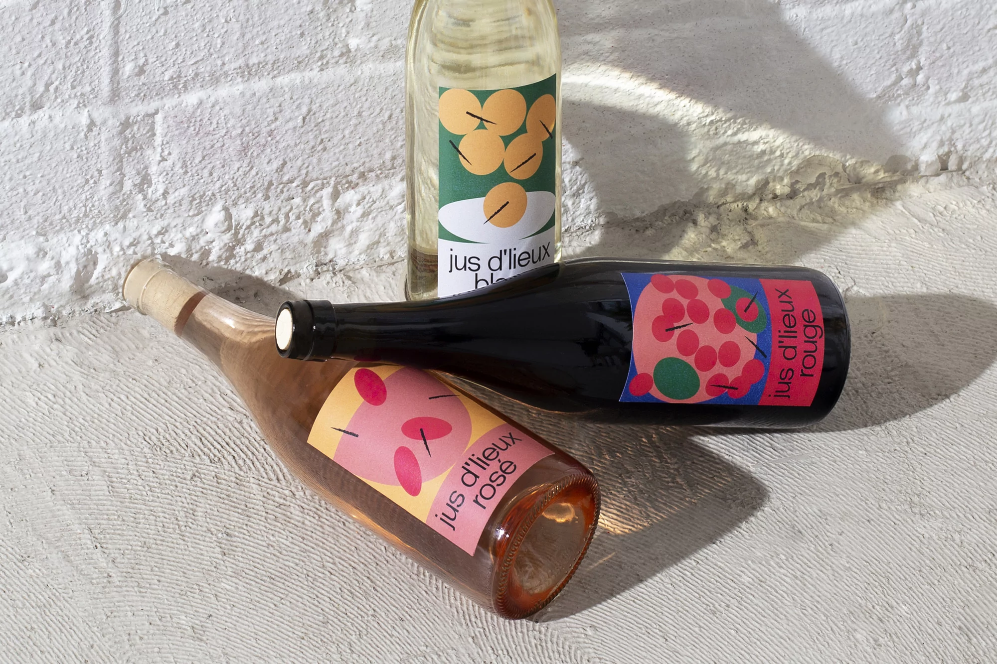 Three bottles of colorful Jus d'Lieux fruit juice, in rose, red, and white varieties, sit together on a textured white surface.