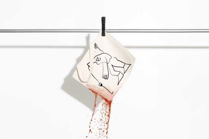 Hanging illustration of figure with blood splatter on white cloth, attached with a clothespin on metal bar against white backdrop.