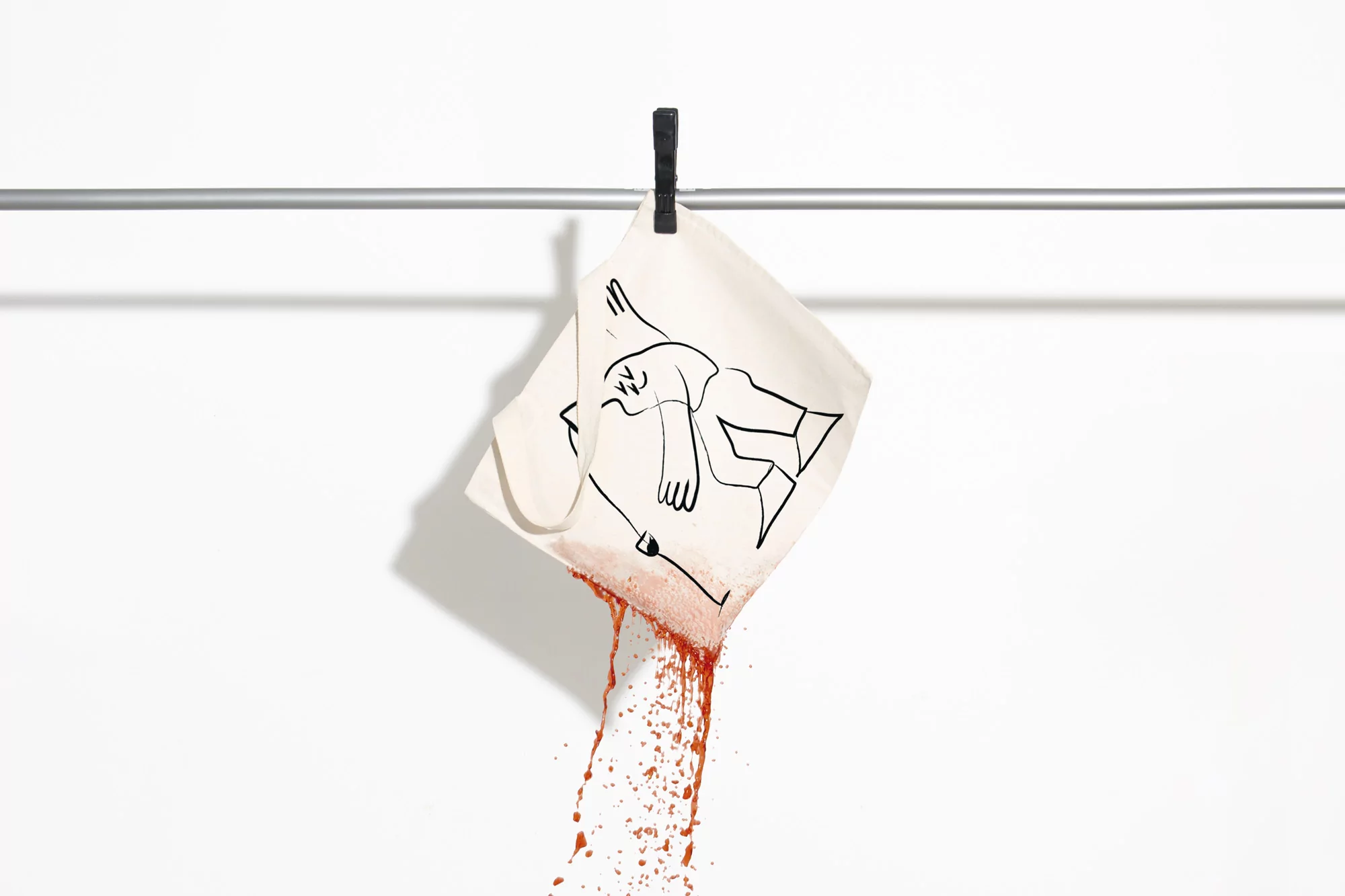 Canvas tote bag hanging from a metal rod with a black clip, leaking red liquid from its lower corner against white seamless.