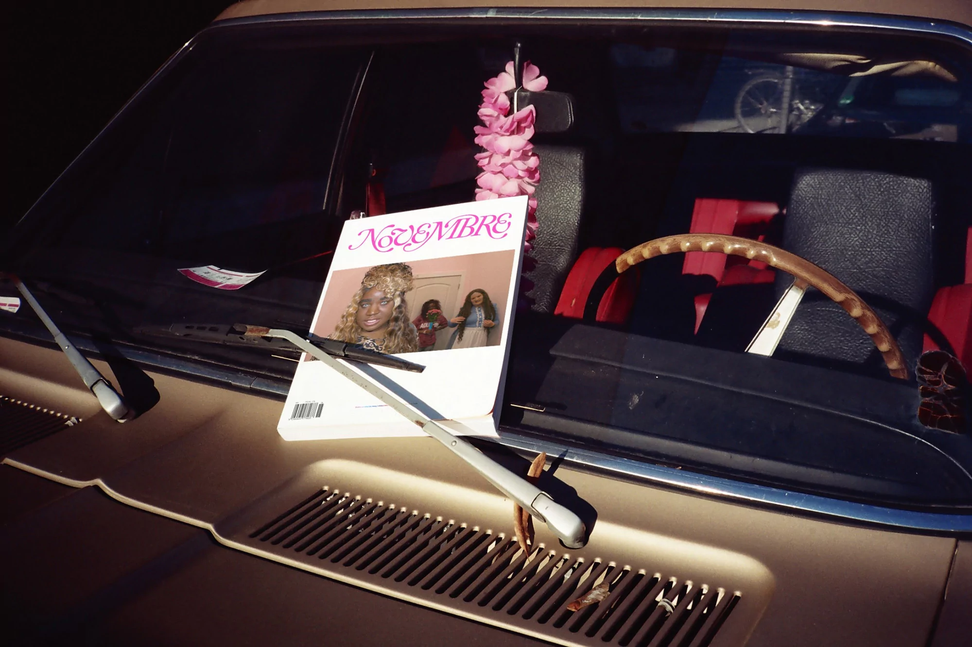 Vintage car hood displays a magazine cover with a portrait of a woman with blonde hair next to a pink floral decoration and black car interior.