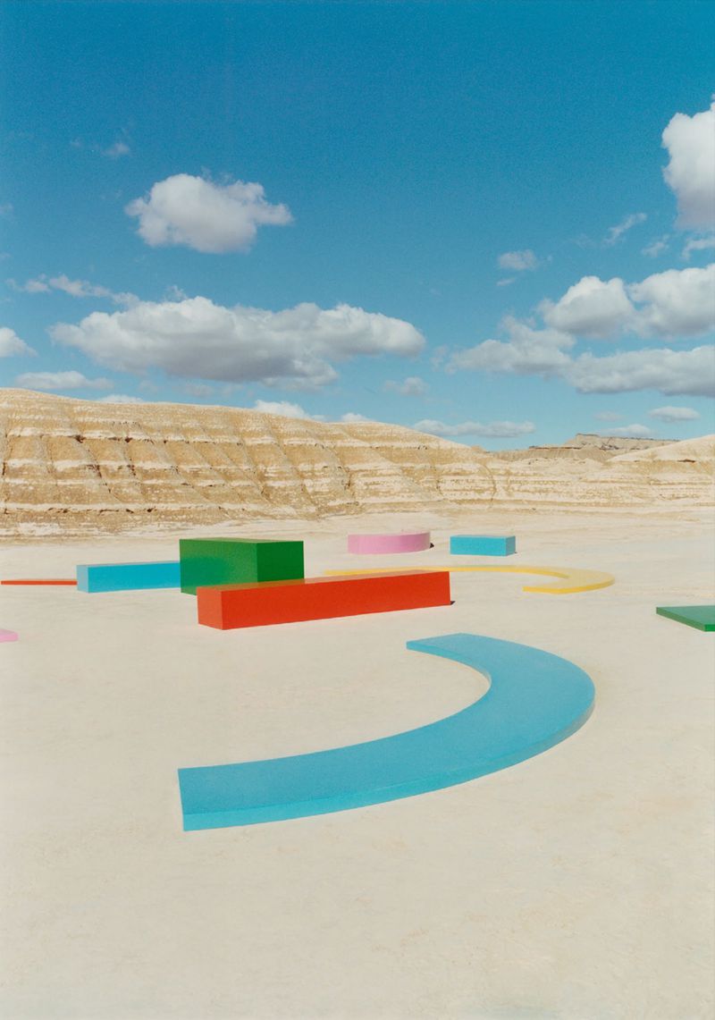 Abstract art installation in desert landscape under blue sky. Geometric shapes, vibrant colors, barren terrain, and clear sky.