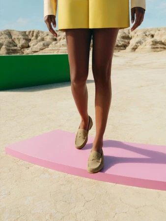 Woman walking in neutral loafers with yellow skirt and long-sleeved top against a desert and geometric set background.