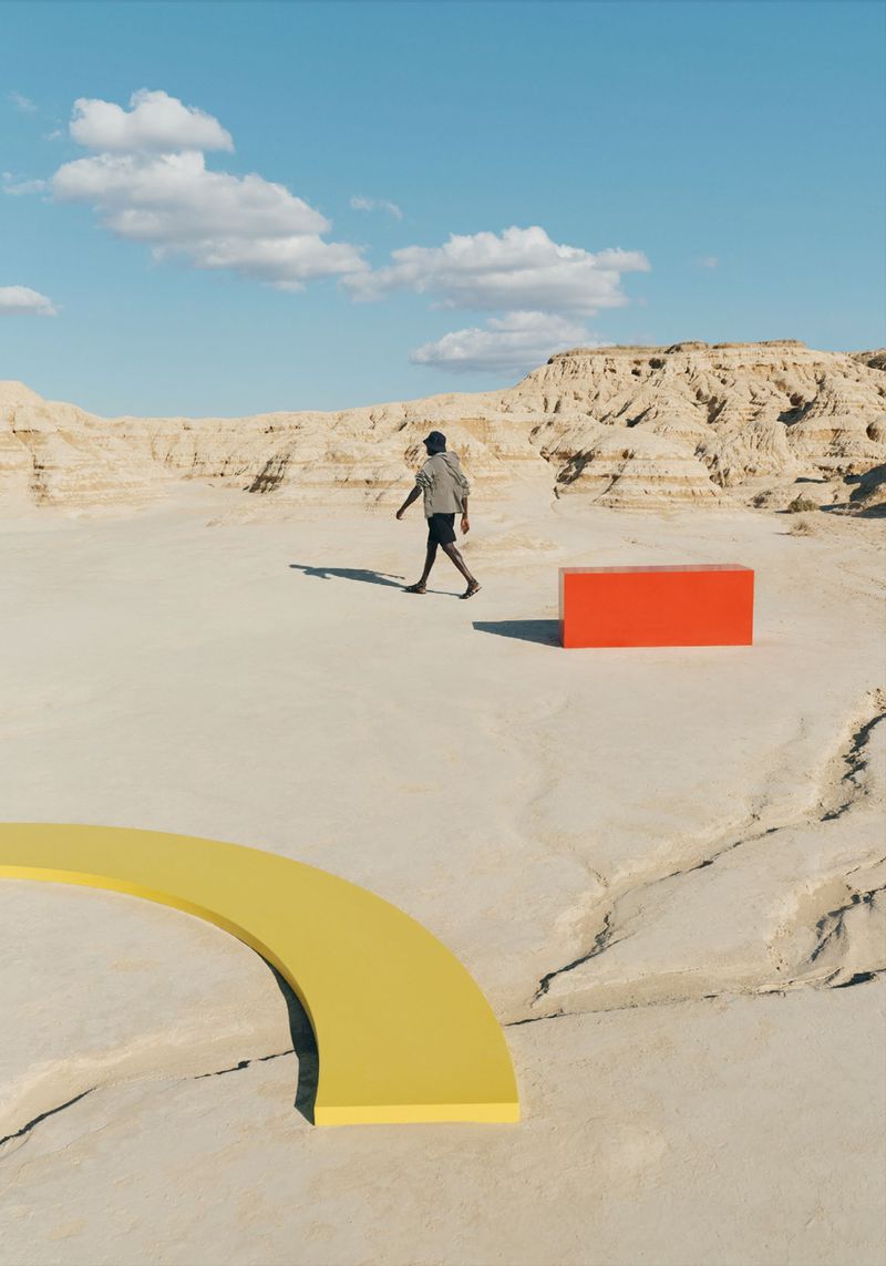 Person walks in surreal desert landscape near geometric shapes against a blue sky with clouds, creating an abstract visual.