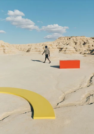 Person walks in surreal desert landscape near geometric shapes against a blue sky with clouds, creating an abstract visual.