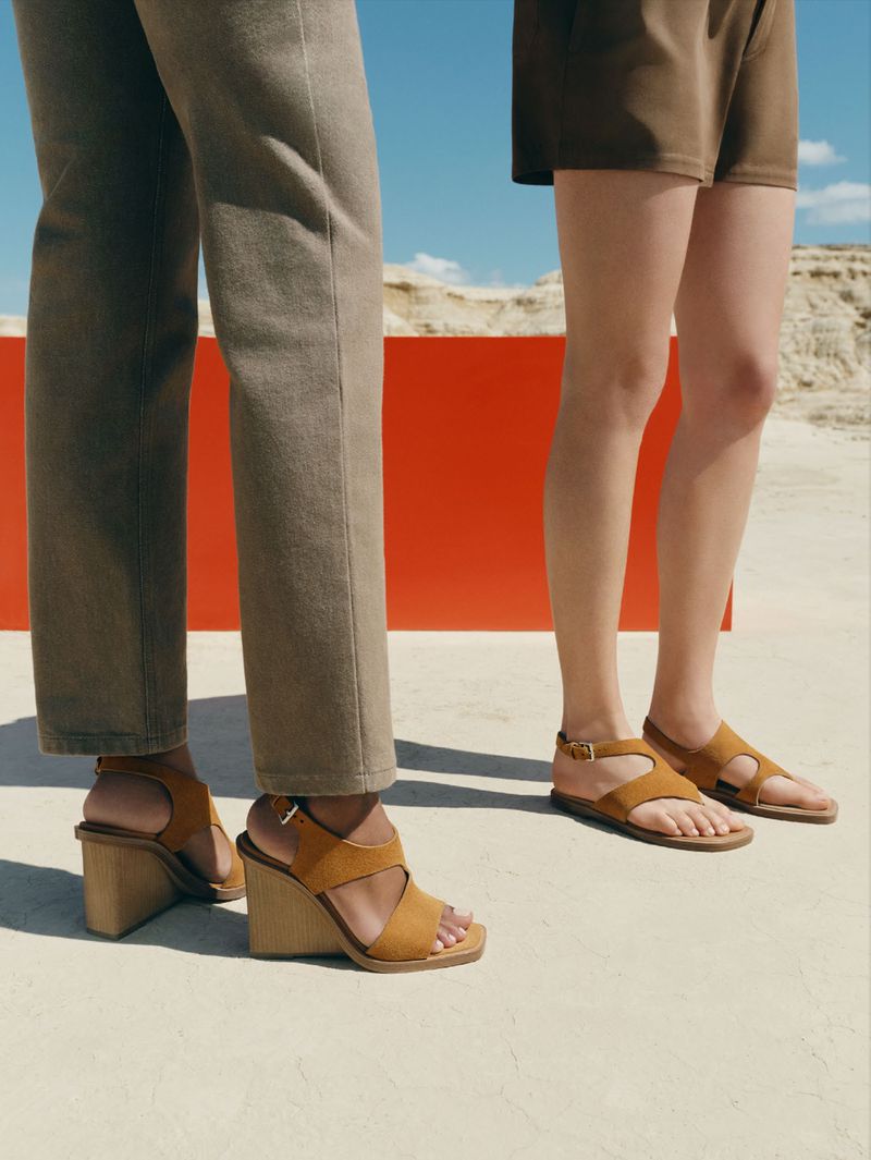 Two women modeling tan-colored leather sandals outdoors against a red accent wall and a desert landscape under a bright blue sky.