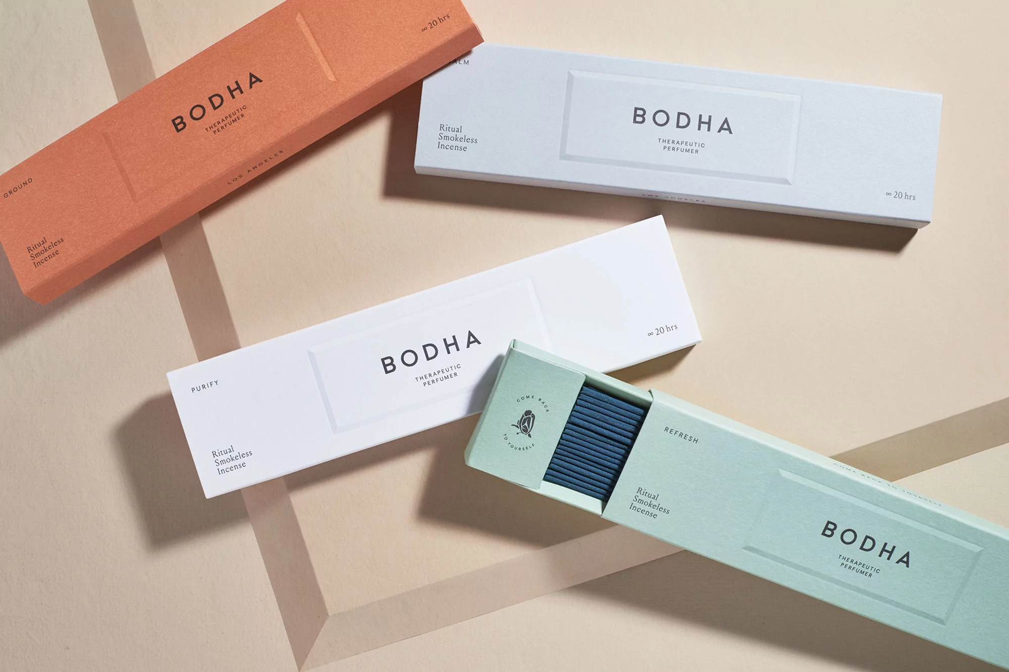 Assortment of Bodha therapeutic perfumes and ritual smokeless incense boxes in peach, white, and sage, arranged on a geometric background.