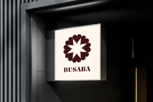 Square illuminated sign displaying the Busaba logo with a dark floral flower emblem on a pale background