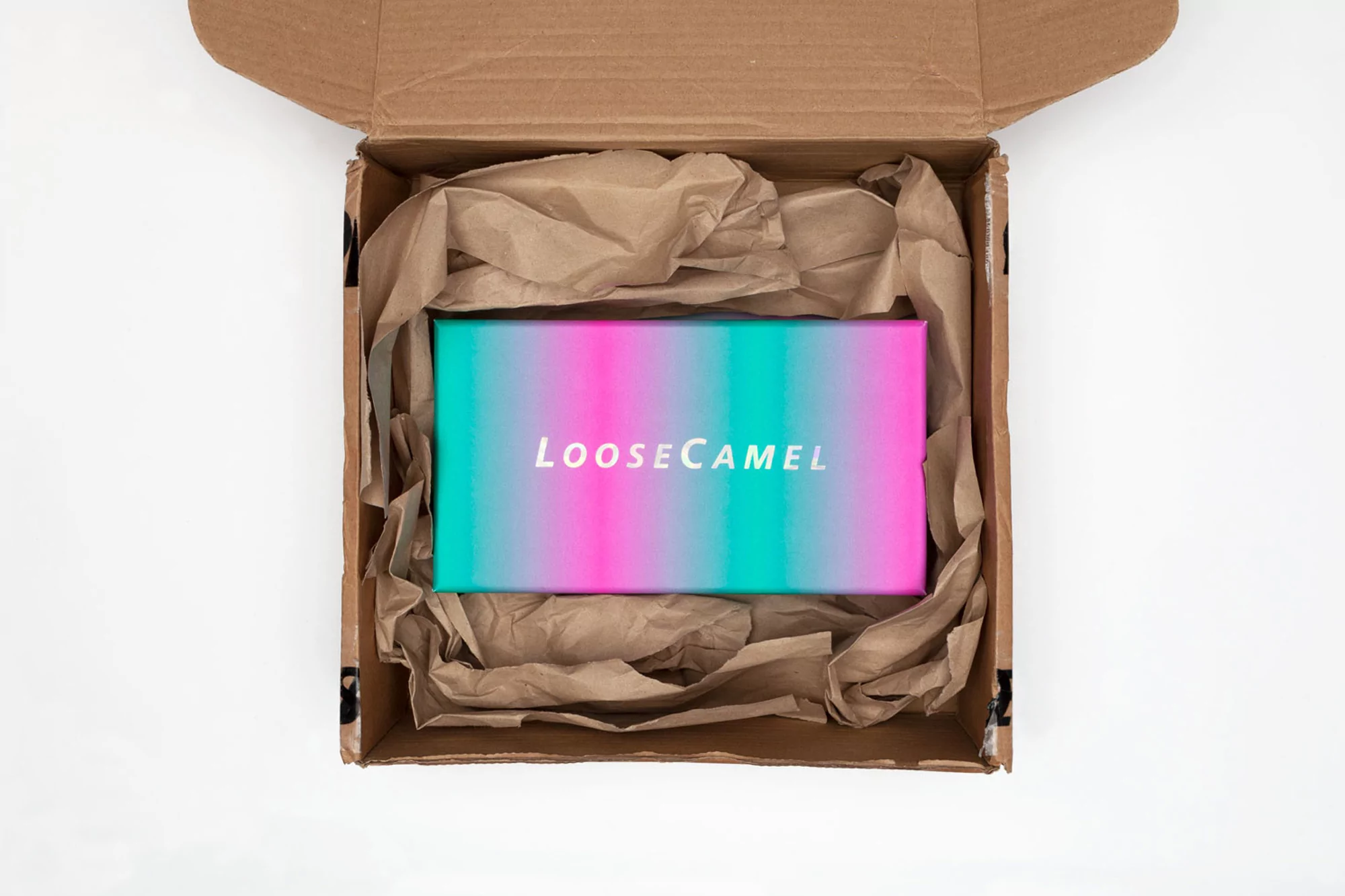 Top-down view of an open cardboard box filled with brown packing paper, revealing a pink and turquoise product box inside.