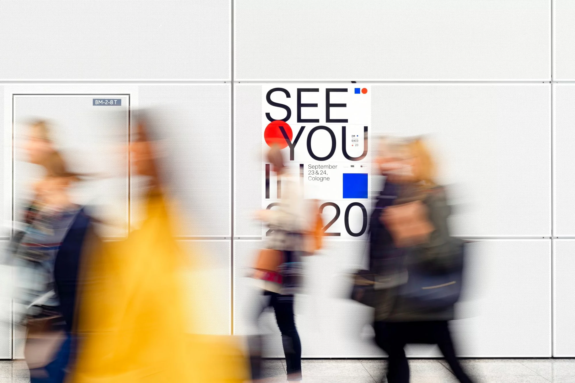 Blurred pedestrians walk past an event poster on a white tiled wall. The event poster is black, red, and blue with a modern design.