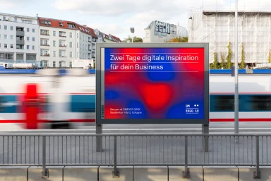 DMEXCO billboard advertisement displaying red and blue graphic patterns on a platform as a commuter train passes in motion blur.