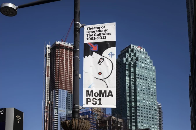 MoMA PS1 art exhibit banner featuring an abstract face attached to a light post in a city environment with skyscrapers.