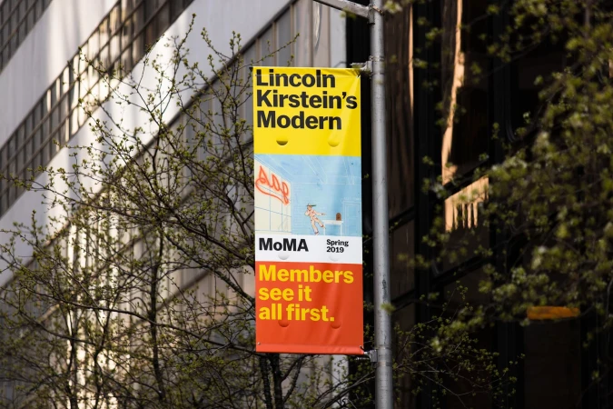 Outdoor banner advertising the Lincoln Kirstein's Modern exhibit at MoMA, featuring an illustration, with a building and trees in the background.