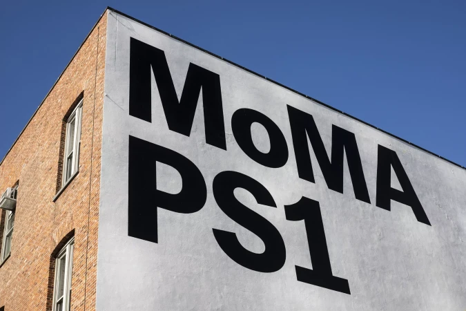Exterior corner of a brick and white building under a blue sky with the large, black block letters of MoMA PS1 displayed on the white facade.